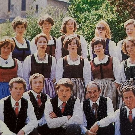 Ein Chor erwachsener Personen in traditioneller deutscher Kleidung posiert vor einer Kirche, lächelt und singt. Im Hintergrund befinden sich Bäume und ein Rasenbereich.