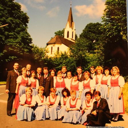 Eine Gruppe von Menschen in traditioneller Kleidung steht vor einer Kirche, mit einem knienden Mann im Vordergrund. Bäume umgeben die Szene.