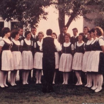 Eine Gruppe von Menschen in traditioneller Kleidung steht in einem Kreis unter einem Baum, wahrscheinlich bei einer Zusammenkunft. Einige Personen stehen hinter ihnen. Im Hintergrund befindet sich ein Gebäude mit Fenstern. Ein mit einem karierten Tuch bedeckter Tisch hat Flaschen darauf.