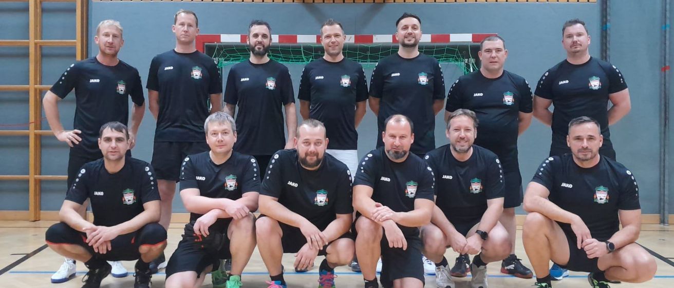 Eine Gruppe von Männern in schwarzen Trikots posiert für ein Foto in einem Fitnessstudio mit einem Basketballkorb im Hintergrund.