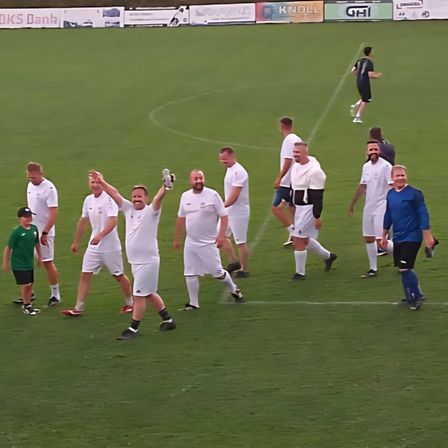 Eine Gruppe von Männern in weißen Trikots läuft auf einem Fußballfeld. Ein Mann hebt die Hände und ein anderer hält einen Ball. Mehrere Spieler gehen zur Mittellinie.