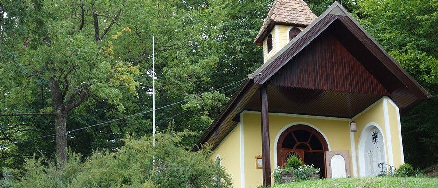 Eine kleine gelbe Kirche mit einem braunen Dach steht auf einem Hügel, umgeben von Bäumen und Sträuchern. Die Kirche hat einen Glockenturm und ein Kreuz auf dem Dach.