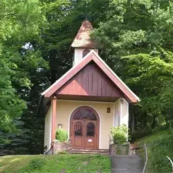 Eine kleine Kapelle mit einer braunen Tür und einem braunen Dach ist von grünen Bäumen und Pflanzen umgeben.