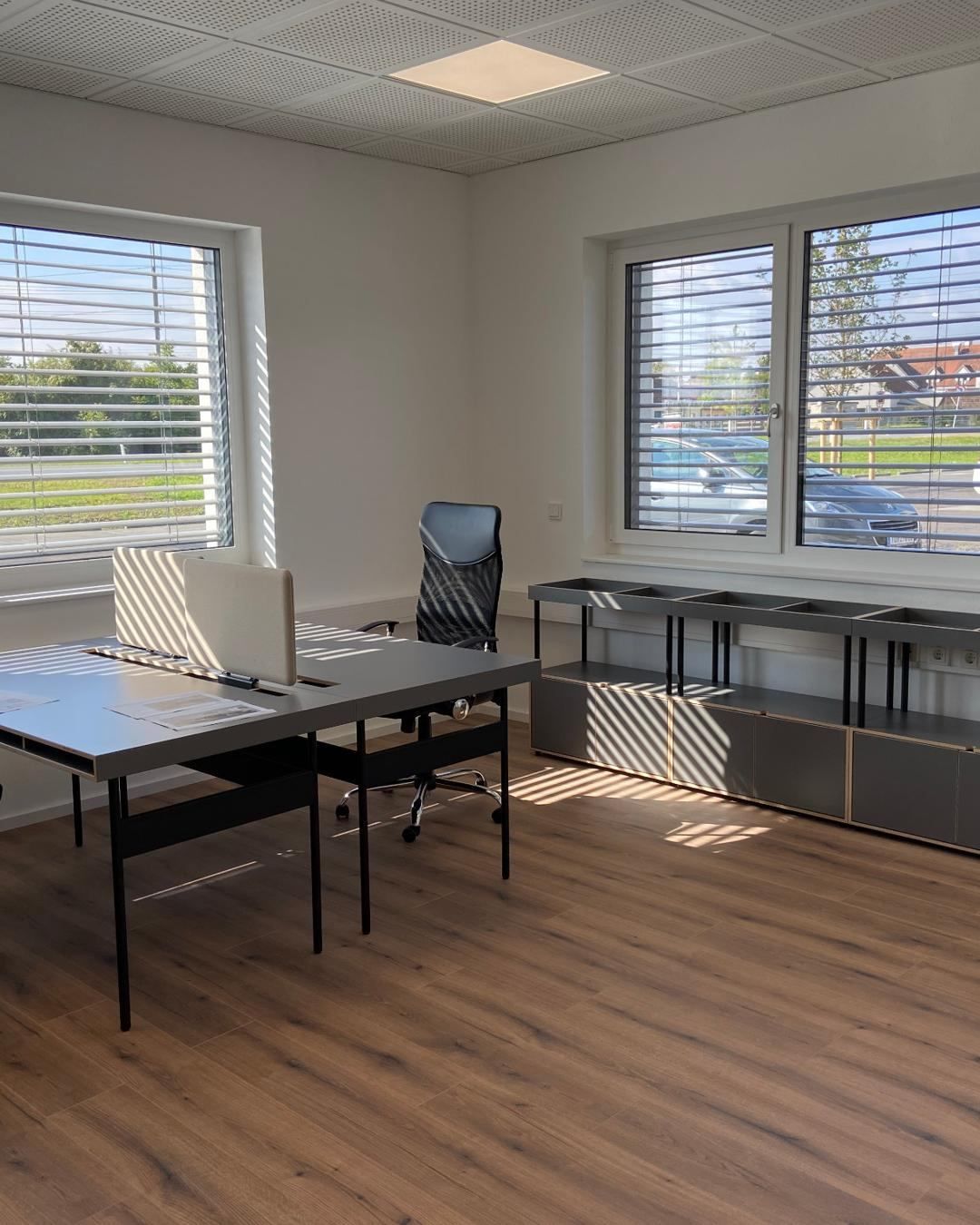 Ein leeres Büro mit einem Schreibtisch, zwei Stühlen und Fenstern mit Jalousien. Vor den Fenstern ist ein Auto neben einem Haus geparkt. Der Boden ist aus Holz.