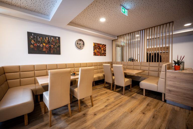 Ein modernes Restaurant mit beigen Ledersitzen und Holztischen. Kunstwerke schmücken die Wände und eine Uhr ist montiert. Ein Topf mit Pflanze steht auf einem Tresen.