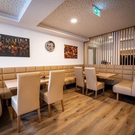Ein modernes Restaurant mit beigen Ledersitzen und Holztischen. Kunstwerke schmücken die Wände und eine Uhr ist montiert. Ein Topf mit Pflanze steht auf einem Tresen.