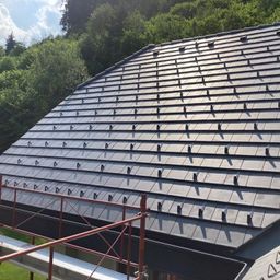 Ein Dach mit geometrischem Fliesenmuster wird gebaut, umgeben von Bäumen und Gerüsten an einem sonnigen Tag.