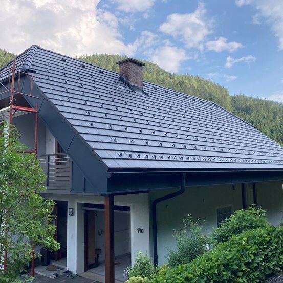 Ein Haus mit einem grauen Schieferdach und Solarpanelen. Das Haus ist von Bäumen und Pflanzen umgeben, mit einem Berg im Hintergrund.