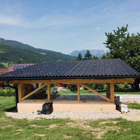 Ein kleines Holzhaus mit schwarzem Dach und Solarmodulen. Es ist von Bäumen und Gras umgeben, mit Bergen im Hintergrund.