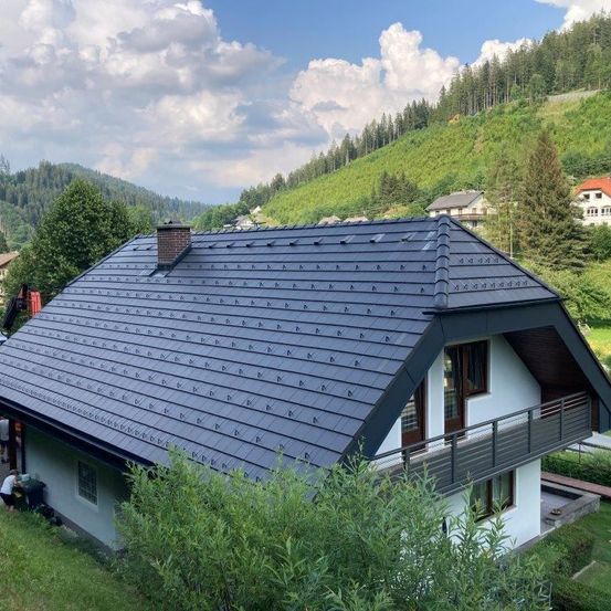 Ein Haus mit grauem Dach, Solarpanelen und einem Schornstein steht auf einem Hügel mit einem grünen Hang. Bäume und Büsche umgeben das Haus. Der Himmel ist blau mit weißen Wolken.