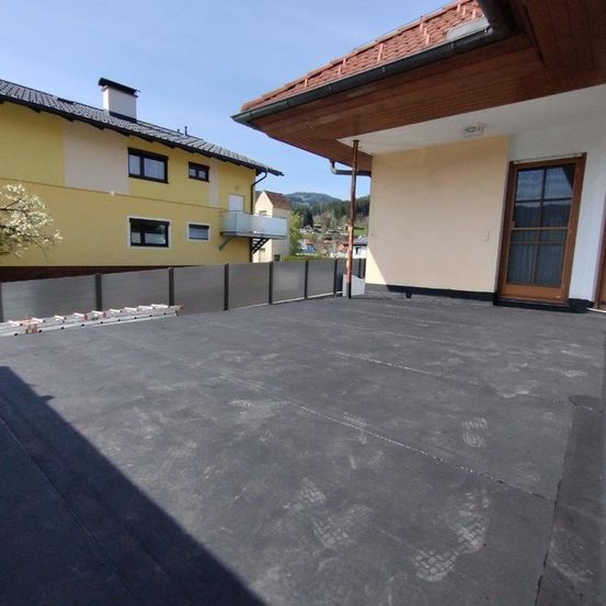Ein Dach mit einem neuen schwarzen Dach. Ein gelbes Haus mit Schornstein befindet sich daneben. Eine Metallzaun und eine Holztür sind sichtbar. Das Dach hat Fußabdrücke.