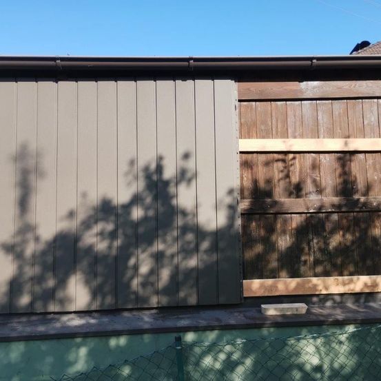 Eine graue Metallverkleidung neben einem hölzernen Zaun mit Schatten, der von Bäumen geworfen wird. Der Zaun ist teilweise geöffnet und eine Holzplatte ruht auf der Schwelle.