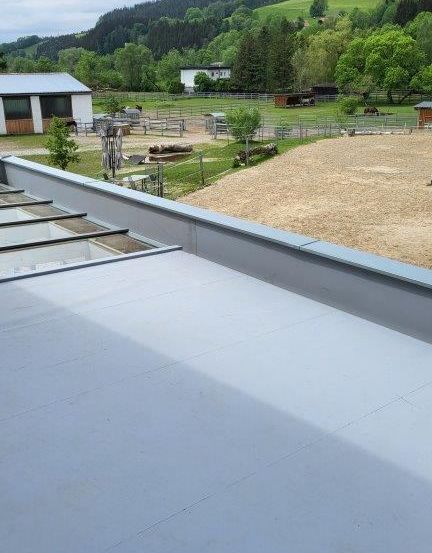 Ein Dach mit grauem Material, wahrscheinlich einer wasserdichten Membran, überblickt einen Bauernhof mit eingezäunten Bereichen und Bäumen. Im Hintergrund gibt es Hügel und einen bewölkten Himmel.