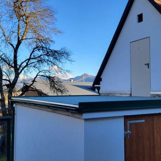 Ein weißes Gebäude mit einem braunen Dach und einer Holztür befindet sich neben einem Baum. Ein kleines Gebäude steht vor dem Baum. Der Himmel ist klar mit einigen Wolken.