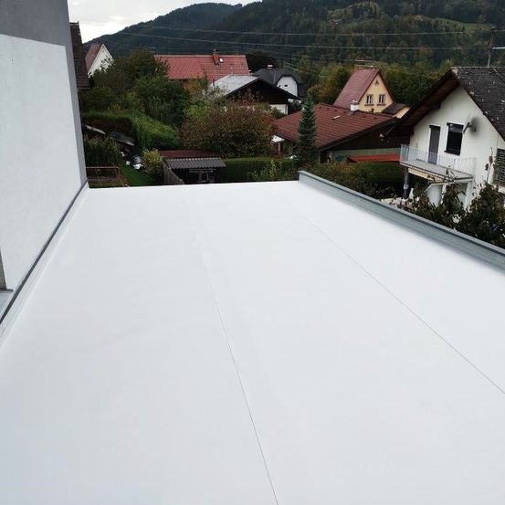 Ein weißes Flachdach mit Dachrinne in einer Stadt mit Häusern und Grünflächen. Im Hintergrund sind Berge.