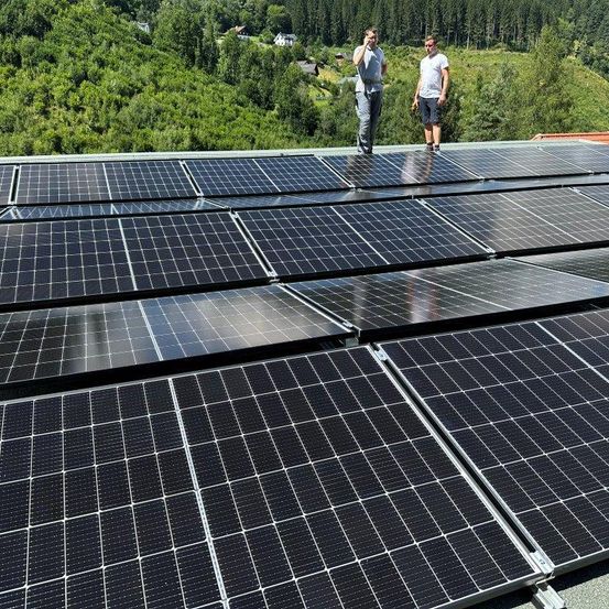 Zwei Männer stehen auf einem mit Solarmodulen bedeckten Dach. Ein Mann hält ein Werkzeug, während der andere telefoniert. Bäume und Häuser sind im Hintergrund zu sehen.