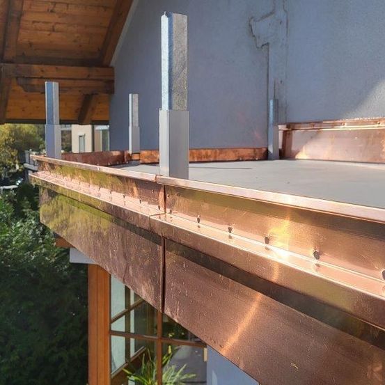 Ein kupfergedeckter Balkon eines Hauses mit grauer Wand, metallischen Pfosten und einem hölzernen Fensterladen. Bäume stehen hinter dem Balkon.