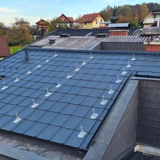 Luftaufnahme eines Hauses mit einem blauen Dach, Solarmodulen und Schornsteinen. Das Dach hat mehrere weiße Solarmontagesysteme.