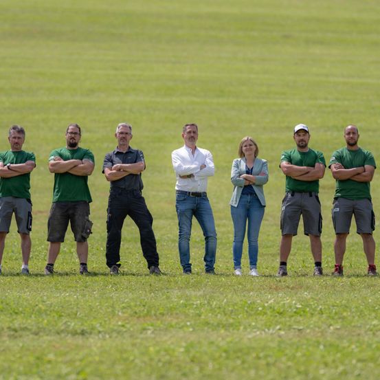 Sieben Menschen stehen in einer Reihe in einem grünen Feld, alle in grünen Hemden und Shorts. Der Mann in der Mitte trägt ein weißes Hemd und blaue Jeans. Die Frau trägt eine leichte Jacke und Jeans. Sie haben alle ihre Arme gefaltet.