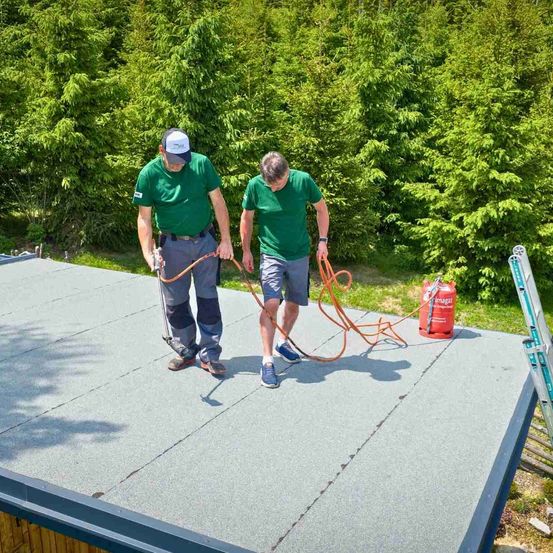 Zwei Männer in grünen Shirts und Shorts arbeiten auf einem Dach und halten einen Schlauch und ein Werkzeug. Dahinter sind ein roter Tank und eine Leiter zu sehen, mit grünen Büschen im Hintergrund.