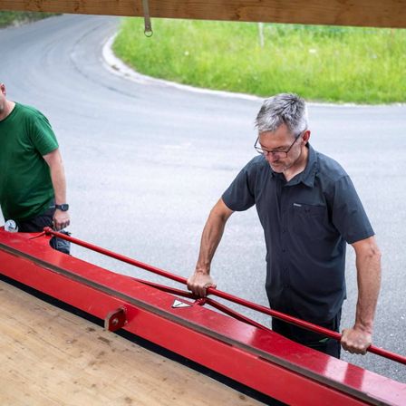 Zwei Männer arbeiten an einer roten Maschine auf der Straße. Einer trägt eine Brille und hält einen roten Griff.