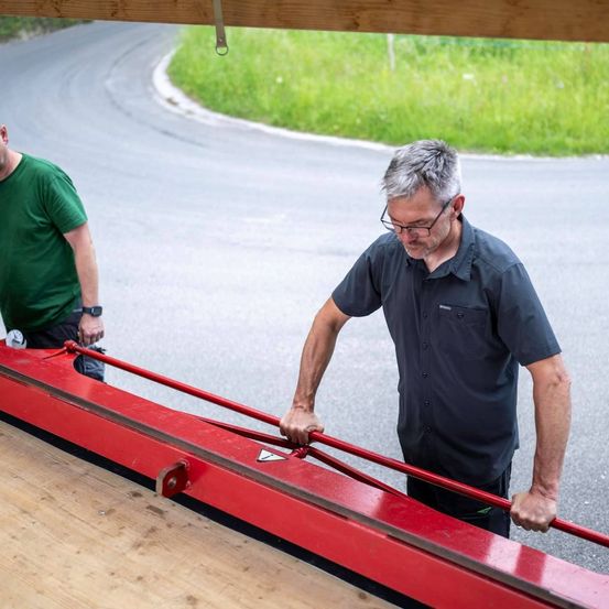 Zwei Männer arbeiten an einer roten Maschine auf der Straße. Einer trägt eine Brille und hält einen roten Griff.