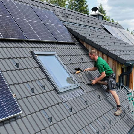 Ein Mann in einem grünen Shirt und Shorts installiert Solarmodule auf einem grauen Dach.