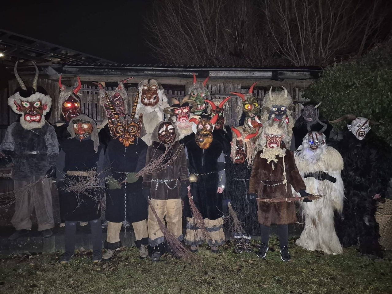 Eine Gruppe von Menschen in aufwendigen Teufelskostümen mit roten Hörnern und Masken steht bei Nacht draußen. Sie halten Stöcke in den Händen und einige tragen Handschuhe. Im Hintergrund ist ein Holzzaun und ein Haus zu sehen.