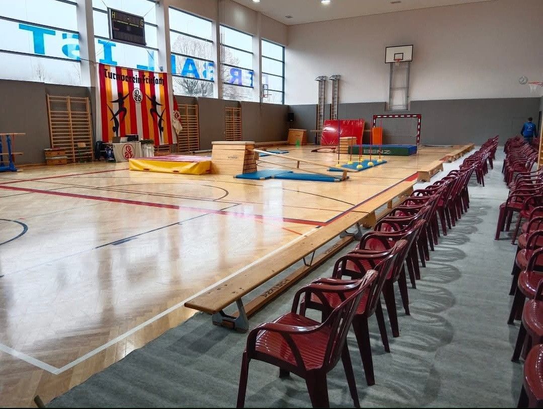 Ein leerer Sportsaal mit Basketballkorb, Turngeräten und Reihen roter Stühle. Es gibt auch Banner und einen Tisch mit einer Topfpflanze.