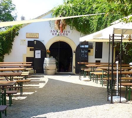 Ein Außenbereich eines Restaurants mit Holztischen und Bänken unter weißen Sonnenschirmen. Das Gebäude hat einen gewölbten Eingang und einen Fass vor der Tür.