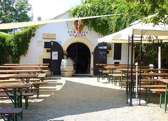 Ein Außenbereich eines Restaurants mit Holztischen und Bänken unter weißen Sonnenschirmen. Das Gebäude hat einen gewölbten Eingang und einen Fass vor der Tür.