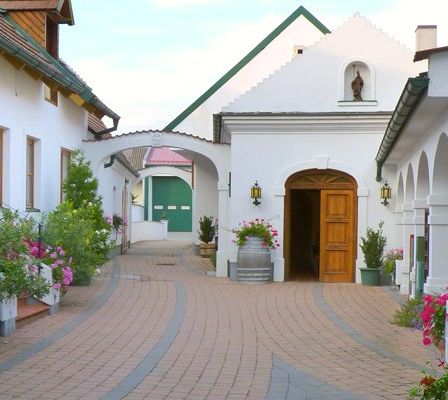 Ein gepflasterter Weg führt zu einer Holztür in einem Innenhof, umgeben von weißen Gebäuden, üppiger Grünfläche und bunten Blumen.