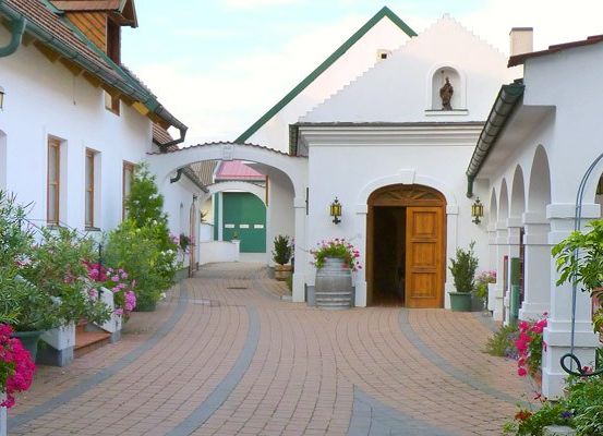 Ein gepflasterter Weg führt zu einer Holztür in einem Innenhof, umgeben von weißen Gebäuden, üppiger Grünfläche und bunten Blumen.