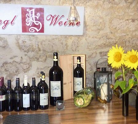 Eine Steinmauer zeigt ein Schild mit der Aufschrift 'Siegl Weine'. Ein Holztisch hält verschiedene Weinflaschen, Gläser und Sonnenblumen in schwarzen Vasen. Eine Laterne und eine Kerze stehen ebenfalls auf dem Tisch.
