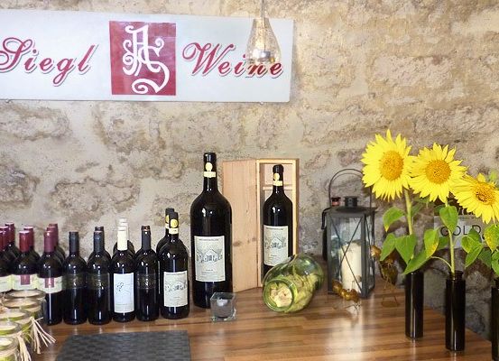 Eine Steinmauer zeigt ein Schild mit der Aufschrift 'Siegl Weine'. Ein Holztisch hält verschiedene Weinflaschen, Gläser und Sonnenblumen in schwarzen Vasen. Eine Laterne und eine Kerze stehen ebenfalls auf dem Tisch.