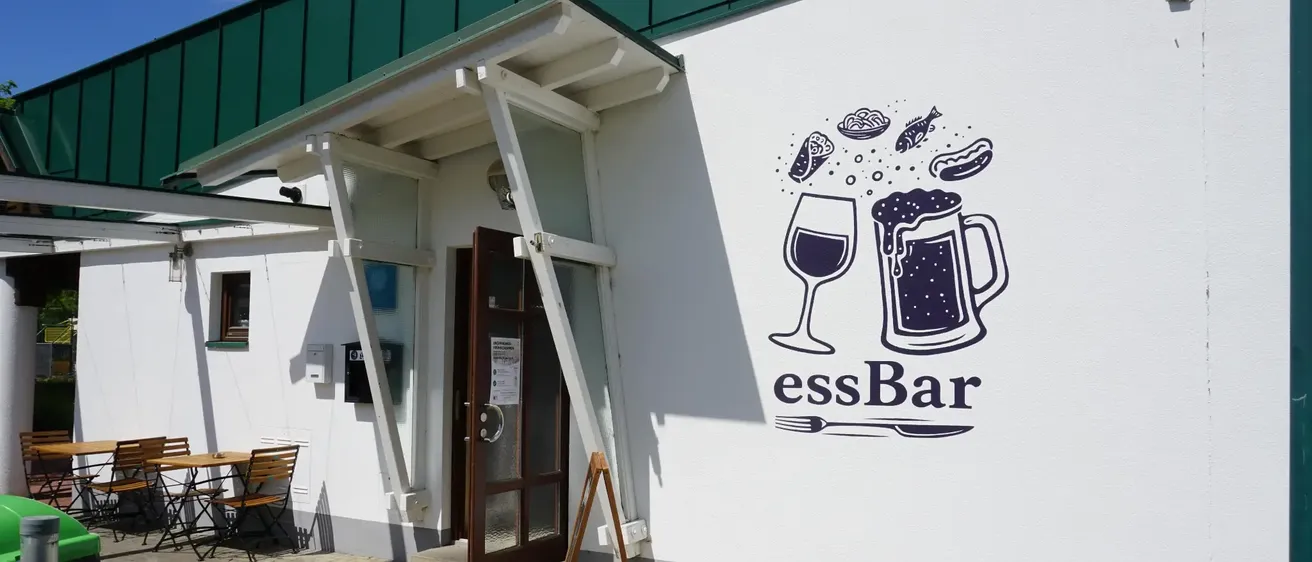 Ein weißes Gebäude mit grünem Dach hat eine Wandmalerei mit einem Bierkrug, einem Weinglas und Essensartikeln. Darunter steht das Wort 'essBar'.