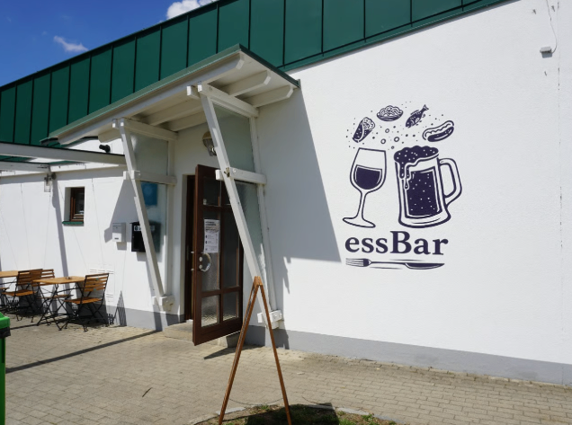 Ein weißes Gebäude mit einem grünen Dach und einer Wandmalerei eines Bierkrugs und eines Weinglases. Darunter steht der Text 'essBar'. Draußen stehen Tische und Stühle.