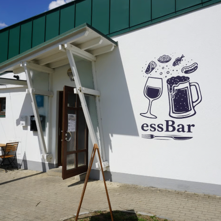 Ein weißes Gebäude mit einem grünen Dach und einer Wandmalerei eines Bierkrugs und eines Weinglases. Darunter steht der Text 'essBar'. Draußen stehen Tische und Stühle.