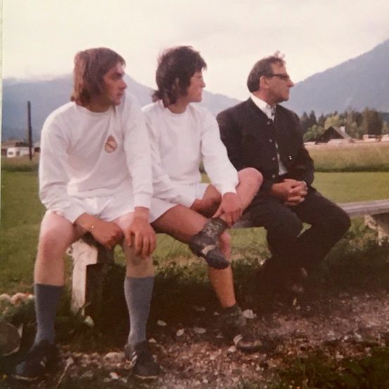 Drei Männer sitzen auf einer Bank in einem Grasfeld mit Bergen im Hintergrund. Zwei tragen weiße Trikots, einer hat eine Brille.