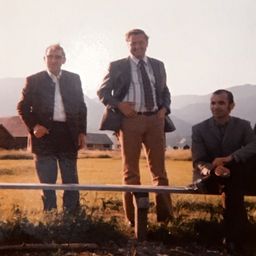 Drei Männer stehen auf einem Feld, zwei stehend und einer sitzend, alle formell gekleidet. Sie stehen vor einem Haus und Bergen im Hintergrund.