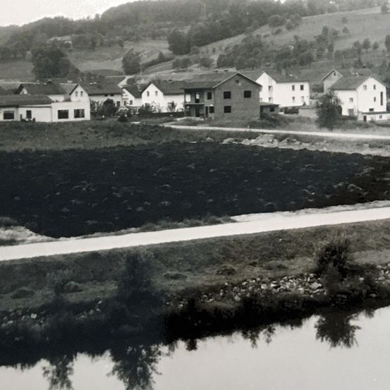 Ein altes Schwarz-Weiß-Foto zeigt ein kleines Dorf mit mehreren Häusern, einem Fluss und einer Straße. Die Häuser liegen eingebettet an einem Hang mit viel Grün und Bäumen im Hintergrund.