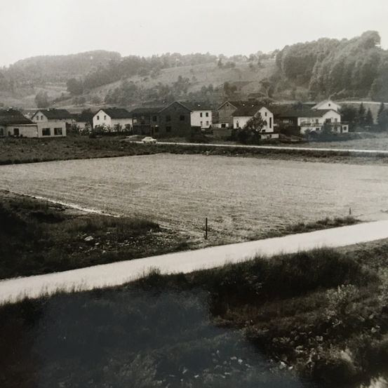 Ein altes Schwarz-Weiß-Foto zeigt eine ländliche Landschaft mit Häusern, einem Feld und einer Straße. Die Häuser sind in einer Reihe, wobei eines in der Mitte etwas größer ist. Im Hintergrund befinden sich Bäume und Hügel. Ein Auto ist in der Nähe geparkt.