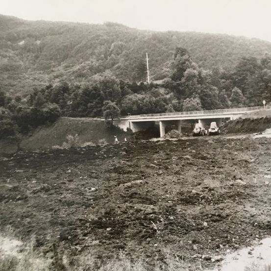 Ein Schwarz-Weiß-Bild, das eine Baustelle zeigt. Fahrzeuge sind darauf abgebildet. Oben befindet sich eine Brücke, im Hintergrund sind Hügel und Bäume zu sehen.