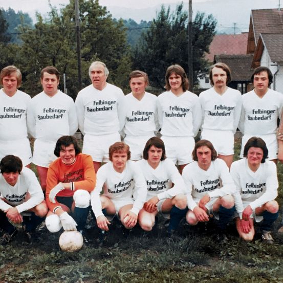 Ein Vintage-Foto einer Fußballmannschaft in weißen Trikots mit 'Felbingerbaudeart' darauf, posiert vor einem Haus mit Bäumen.