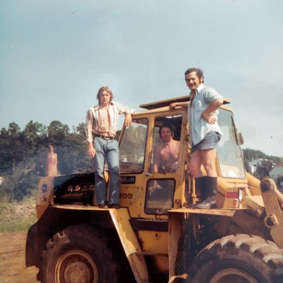 Drei Männer in Freizeitkleidung auf einem Bulldozer, einer steht vorne, einer drinnen und einer lehnt an der Tür. Bäume und Häuser im Hintergrund.