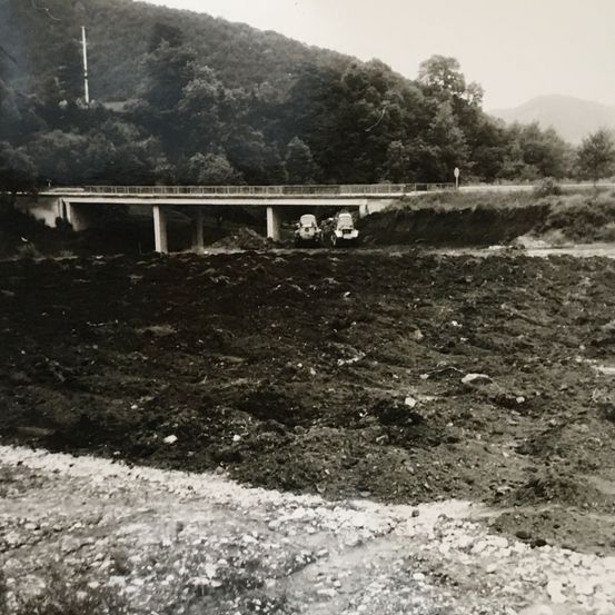 Ein Schwarz-Weiß-Foto zeigt eine Baustelle mit einem im Bau befindlichen Brückenbau und geparkten Fahrzeugen, mit einem bergigen Hintergrund.