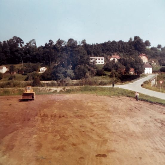 Eine Luftaufnahme einer Baustelle mit einem gelben Bulldozer im Vordergrund. Im Hintergrund befinden sich eine Straße und mehrere Häuser.