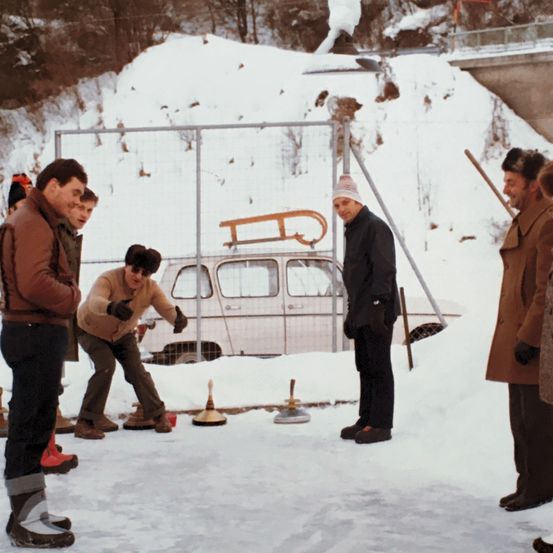 Eine Gruppe von Menschen spielt eine Partie Curling auf einem schneebedeckten Feld. Ein weißes Auto steht hinter einem Metallzaun.