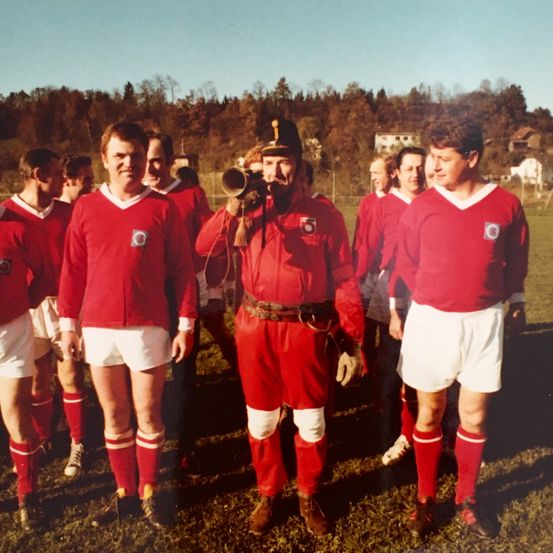 Ein altes Foto einer Fußballmannschaft in roten Trikots, ein Spieler bläst eine Hupe, steht auf einem Feld mit Bäumen und Häusern im Hintergrund.