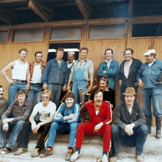 Eine Gruppe von Menschen steht und sitzt vor einem Holzgebäude. Sie tragen Freizeitkleidung und einige haben Hüte. Der Mann im roten Anzug lächelt.